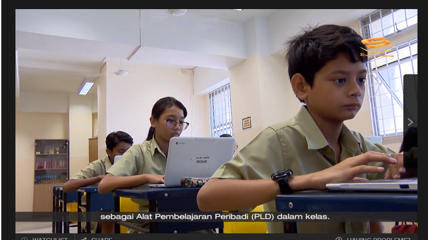 Three students in uniform sit at school desks using laptops, with Malay subtitles at the bottom.